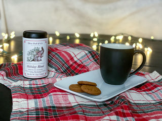 black tea tin and black mug with ginger cookies sitting on red and green plaid blanket outside with Christmas lights under a tent