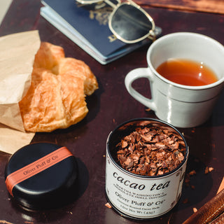 pastry, open tea tin with cacao, half full cup fo tea and sunglasses sitting on wooden table