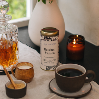 scenic photo of coffee tin, brown mug of coffee, bourbon, a light candle and a vase of flowers on a table