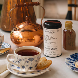 A copper teapot, Colonial Bohea tea canister, honey bottle, and a blue floral tea cup with tea and a biscuit on a saucer.