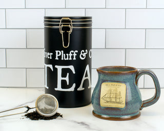 black latch tin and ceramic mug and mesh tea strainer on counter