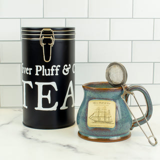 black latch tin and ceramic mug and mesh tea strainer on counter