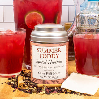 silver tea tin with glasses of red tea and a glass pitcher behind. Cinnamon Sticks, Hibiscus Petals, Ginger, All Spice sprinkled in front, on wooden table