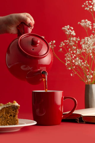 scenic photo of red tea pot pouring tea into red mug beside a slice of carrot cake and white flowers