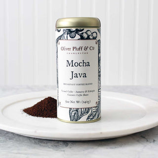 gold coffee tin with black and white flowery images sitting beside a small pile of ground coffee on white plate